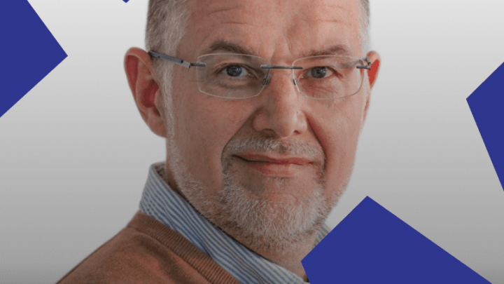 A middle-aged man with glasses and a light beard smiles, wearing a collared shirt and brown sweater. Connected jobsite theme background.