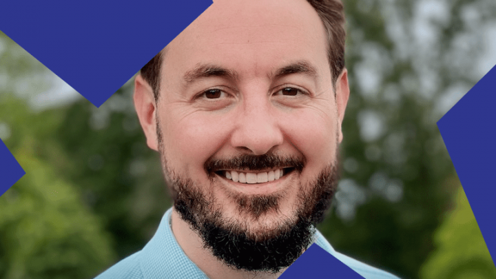 A smiling man with short brown hair and a beard stands outdoors before blurred greenery, with blue geometric shapes overlaying the image, suggesting connected jobsite.