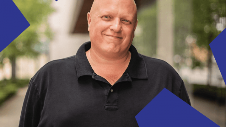 A smiling bald man in a black polo shirt stands outside near trees and glass buildings, partly covered by blue geometric shapes, with construction IoT.