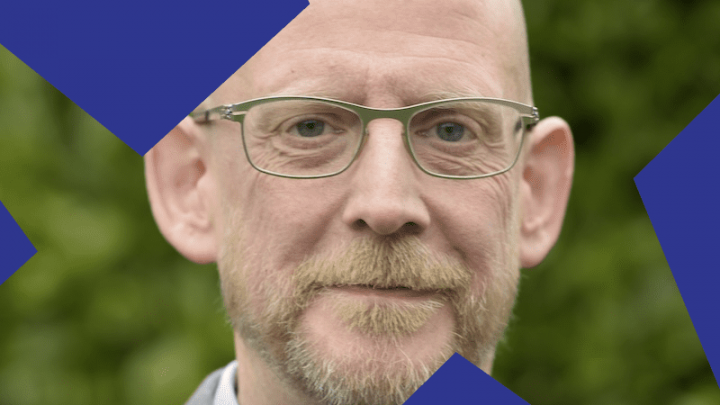 A bald man with glasses and a trimmed beard stands outdoors with greenery behind him, blue shapes overlaying his face—construction IoT.