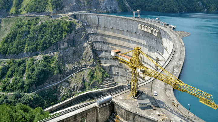 A curved concrete dam spans a river with green hills, a yellow crane, and buildings on top—highlighting construction IoT.