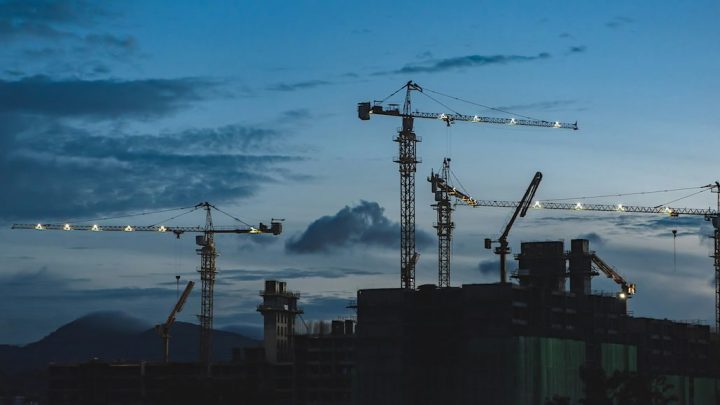 Construction cranes at a dusk jobsite are silhouetted against a blue sky as telematics track their operation in real time.