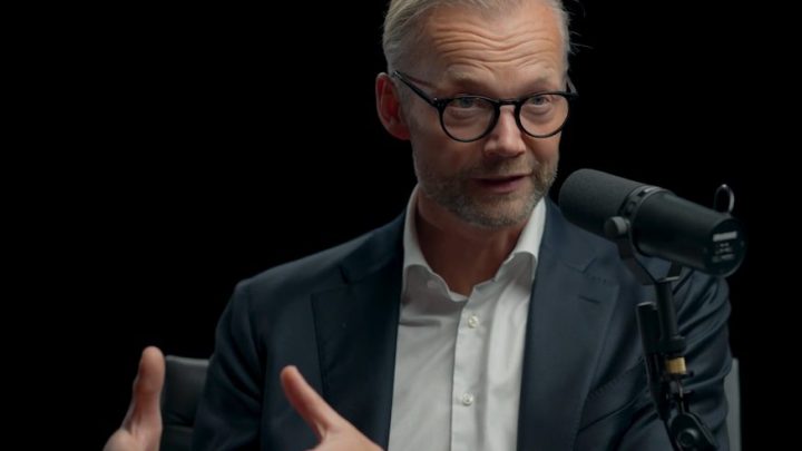 A man in a suit and glasses speaks into a microphone during a podcast, discussing safe equipment access and construction IoT.