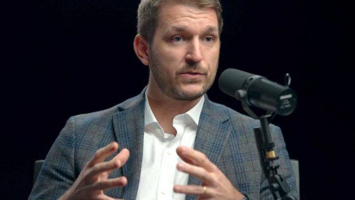 A man in a checked blazer and white shirt speaks into a microphone about safe equipment access in construction fleet management.