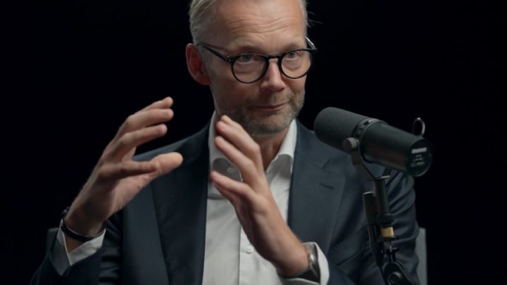 A man in a suit and glasses gestures while speaking into a mic, discussing safe equipment access in a podcast, with a focus on telematics.