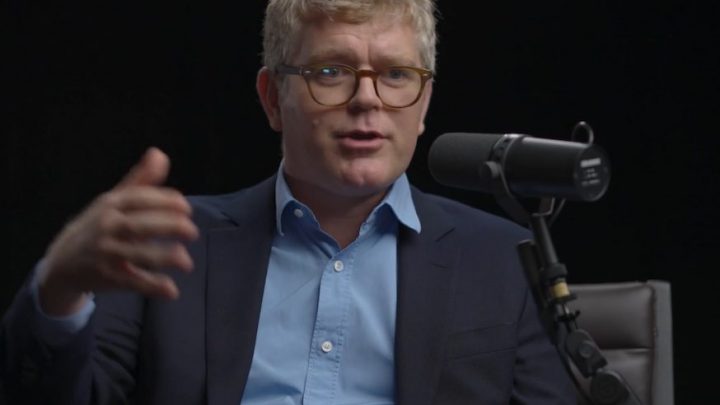 A man in a suit and glasses speaks into a microphone about safe equipment access, likely in a podcast on construction equipment tracking.