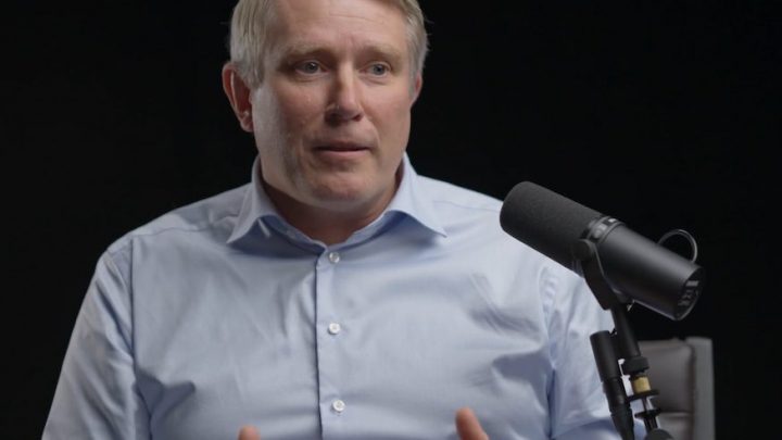 A man in a light blue shirt sits at a microphone, gesturing as he discusses safe equipment access, related to fleet management.
