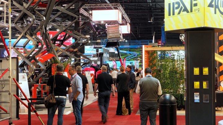 People walk through a busy indoor ire show with industrial equipment, scissor lifts, and booths under bright lights for construction IoT.