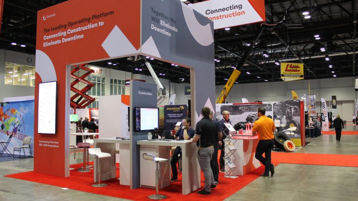 A red and white trade show booth with Trackunit displays, digital screens, and people talking about construction equipment tracking.