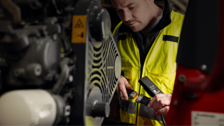 A man in a yellow safety vest inspects machinery with diagnostic equipment at an expo, focused on wires. Construction IoT context.