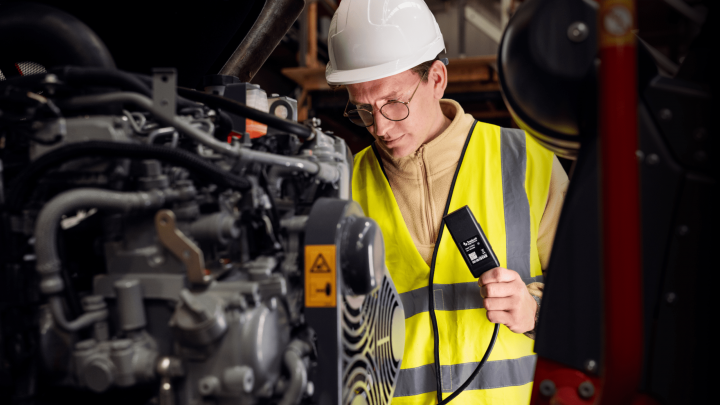 A worker in a hard hat and safety vest inspects machinery with a diagnostic device, supporting fleet management in an industrial setting.