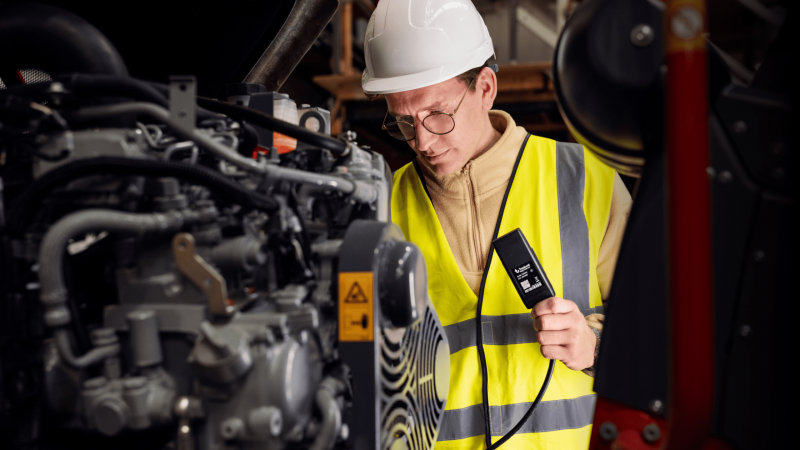 A worker in a hard hat and safety vest inspects machinery with a diagnostic device, supporting fleet management in an industrial setting.
