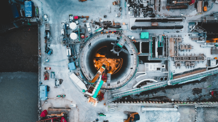 Aerial view of a construction site with a circular tunnel entrance, machinery, and workers; bright lights highlight a connected jobsite.