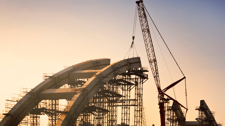 A crane lifts a steel beam onto an arched bridge as project managers oversee progress with construction IoT at sunset.