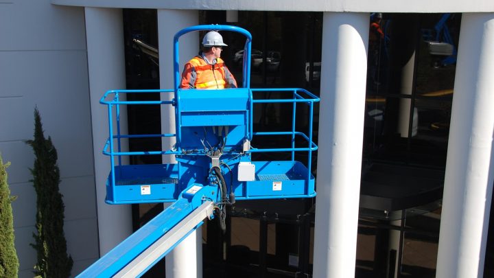 Worker in a hard hat and safety vest operating a blue aerial lift platform near building columns for construction equipment tracking.