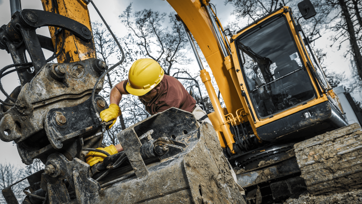 A worker in a yellow hard hat repairs an excavator bucket with aftermarket parts at a muddy site, highlighting construction equipment tracking.