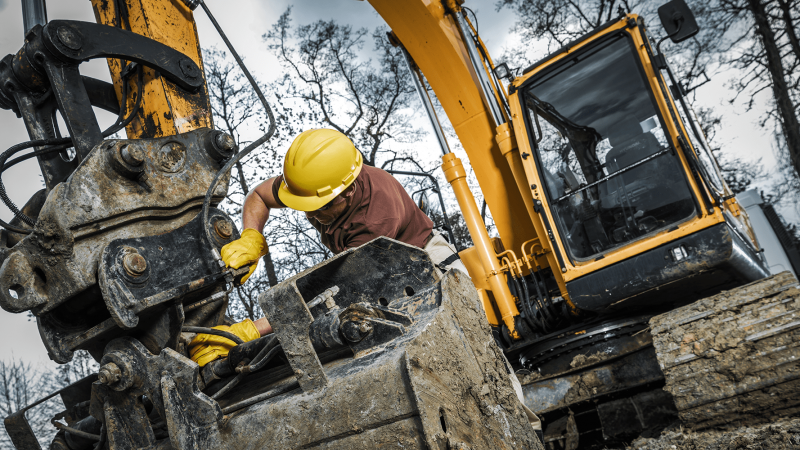 A worker in a yellow hard hat repairs an excavator bucket with aftermarket parts at a muddy site, highlighting construction equipment tracking.