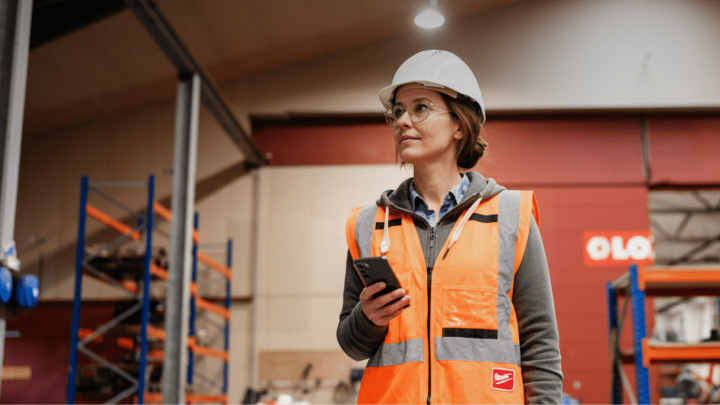 A woman in safety gear oversees installs in a warehouse, using her smartphone on a connected jobsite with industrial shelving behind her.