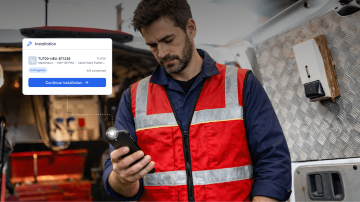 A man in a red safety vest stands by a work van, checking his phone, with an install progress graphic for fleet management.