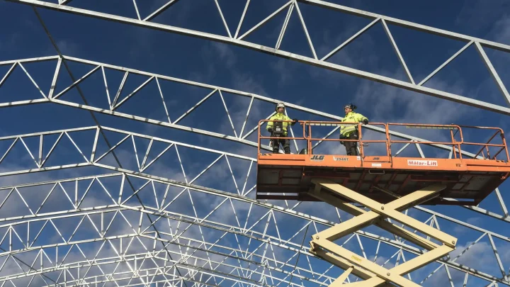 Two workers in high-visibility vests use a MEWP platform to inspect a building’s metal framework on a connected jobsite.