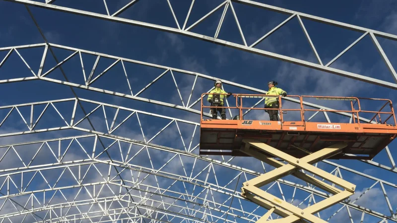 Two workers in high-visibility vests use a MEWP platform to inspect a building’s metal framework on a connected jobsite.