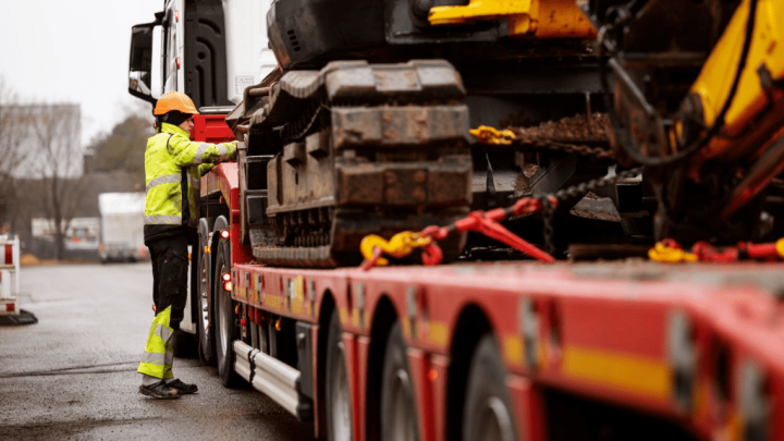 A worker in a yellow vest and helmet checks a red flatbed truck with construction equipment at a connected jobsite.