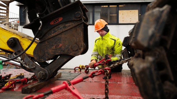 Worker in a yellow safety jacket and orange hard hat secures chains to telematics-enabled construction equipment on a jobsite.