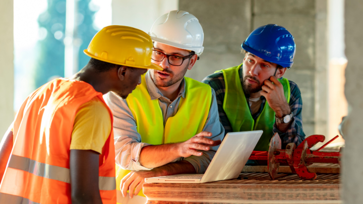 Three construction workers in safety gear look at a laptop on-site, reviewing telematics data for construction equipment tracking.
