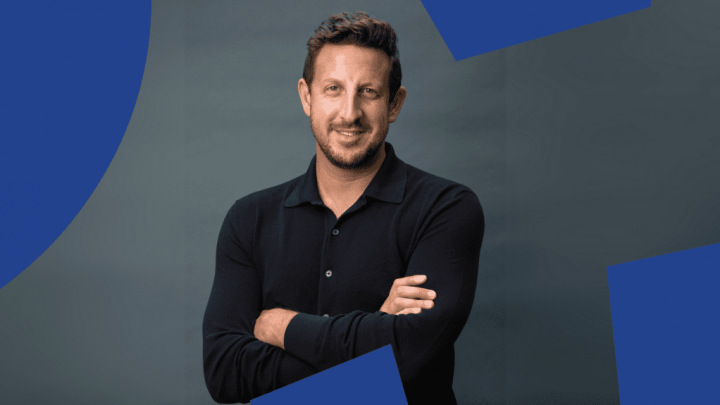 A man with short brown hair and a beard stands smiling, arms crossed, in front of blue telematics-inspired shapes for construction IoT.