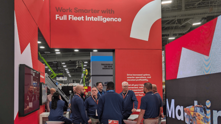 People in blue shirts talk at a trade show booth about an advanced telematics system, with construction equipment tracking displays.