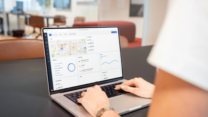 A person uses a laptop showing a telematics dashboard with maps and analytics; office background has chairs and glass walls.