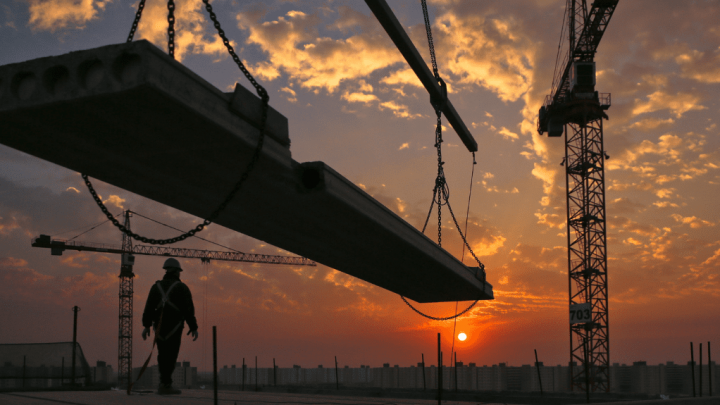 A Gammon Construction worker stands by a suspended concrete beam as cranes operate at sunset on a connected jobsite.