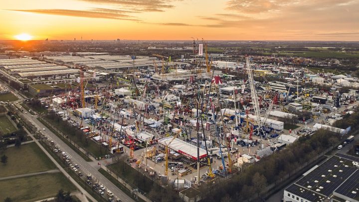 Aerial view of a construction equipment trade show at sunset with cranes, machines, tents, crowds, and fleet management displays.