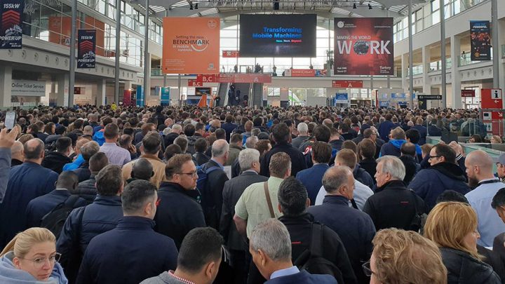A crowd fills the bauma hall, facing a stage with digital screens and banners on downtime reduction in construction equipment tracking.