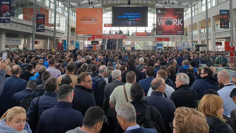 A crowd fills the bauma hall, facing a stage with digital screens and banners on downtime reduction in construction equipment tracking.