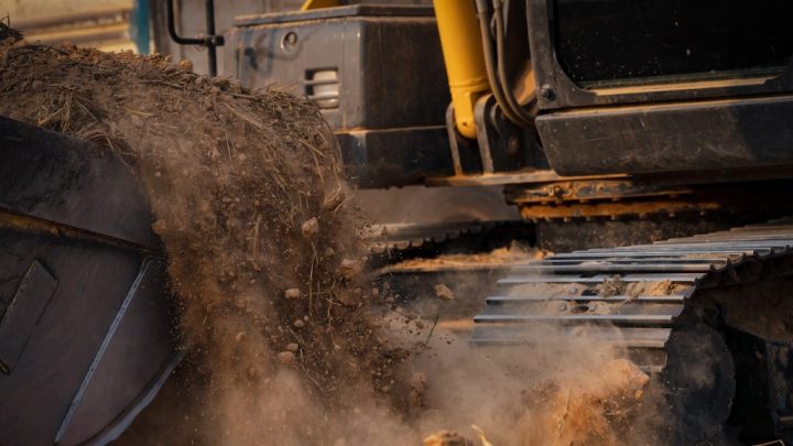 Close-up of a digital excavator dumping dirt onto metal tracks, dust rising at a construction site with connected jobsite technology.