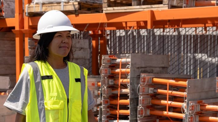 A woman in a hard hat and safety vest stands at a construction site with stacked materials, suggesting fleet management.