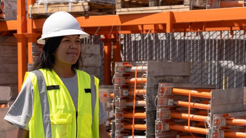 A woman in a hard hat and safety vest stands at a construction site with stacked materials, suggesting fleet management.