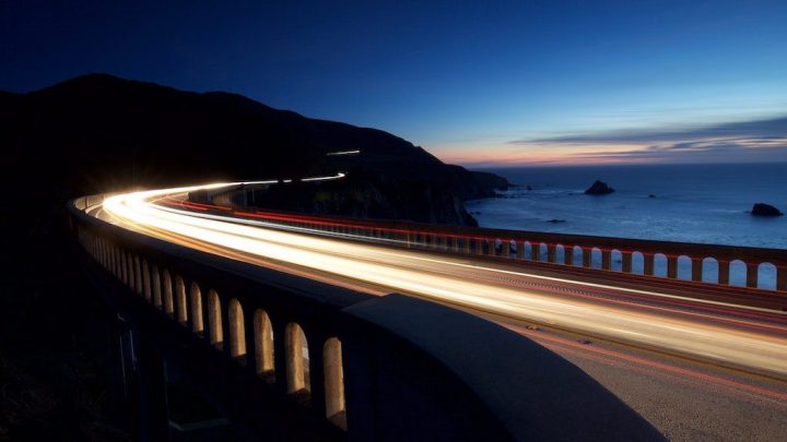 Light trails from cars on a coastal bridge at dusk, ocean and hills behind, highlighting travel speed and fleet management.