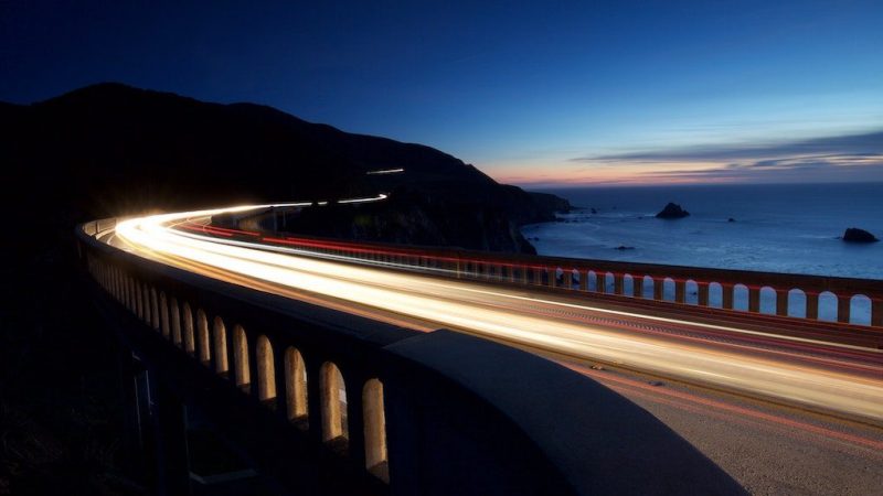 Light trails from cars on a coastal bridge at dusk, ocean and hills behind, highlighting travel speed and fleet management.