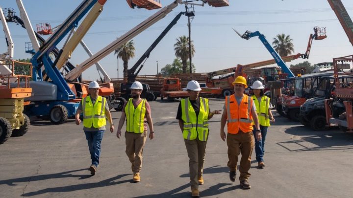 Five construction workers in safety vests and helmets walk through a lot of construction equipment under palm trees, illustrating fleet management.