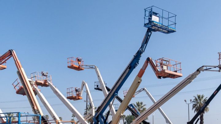 Construction boom lifts with raised platforms extend skyward, safety features visible, palm trees in background; construction fleet management.