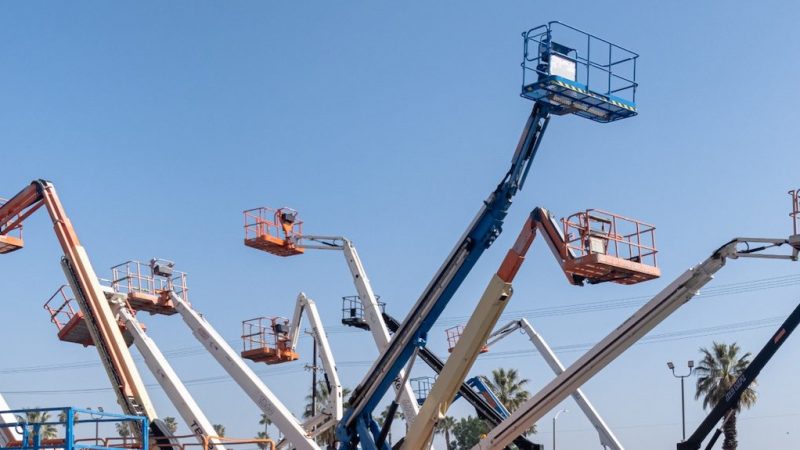 Construction boom lifts with raised platforms extend skyward, safety features visible, palm trees in background; construction fleet management.