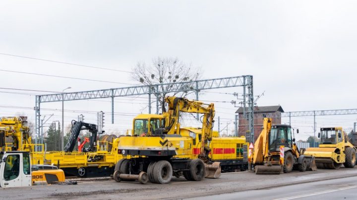 Yellow excavators, bulldozers, and rollers are parked by a road near tracks as workers coordinate construction fleet management.