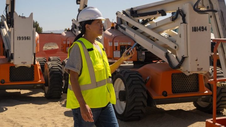 A construction worker in a white helmet and yellow vest uses a walkie-talkie, supervising orange equipment with construction IoT.