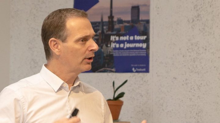 A man in a white shirt gestures as he speaks about reducing downtime, with a poster and potted plant behind him; construction IoT context.