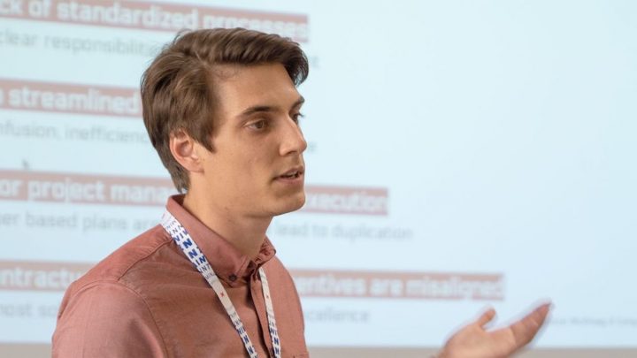 A man in a salmon shirt and lanyard discusses collaboration in construction by a projected slide, highlighting connected jobsite.