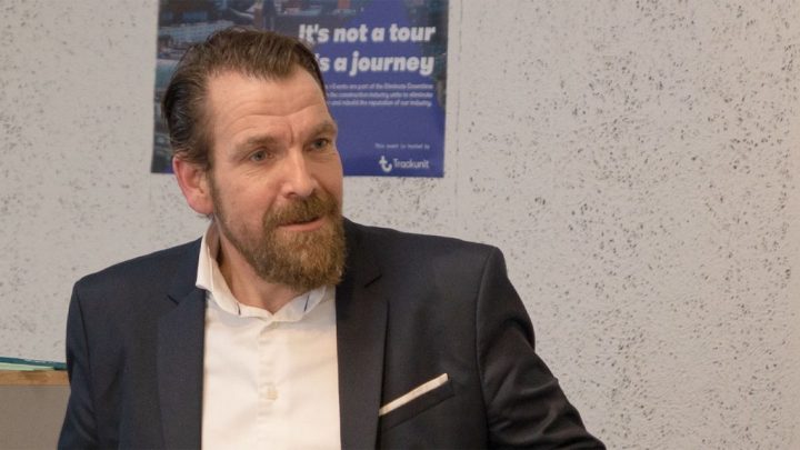 A bearded man in a dark suit and white shirt sits indoors by a textured wall, referencing construction equipment tracking.