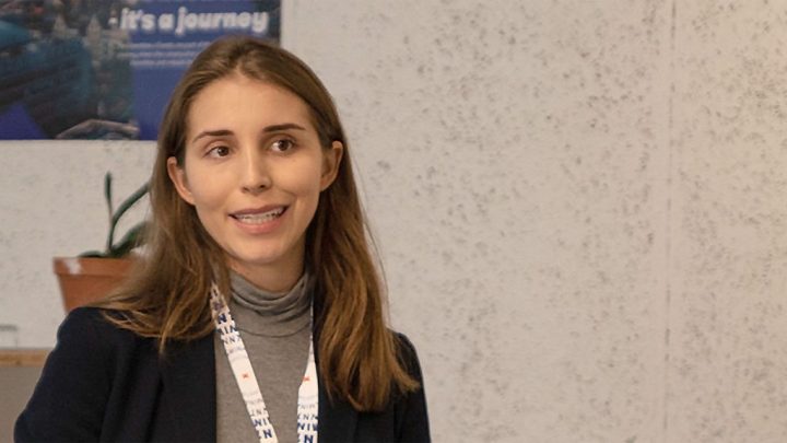 A woman with long brown hair in a blazer and lanyard speaks indoors about construction, with a plant and poster in the background.