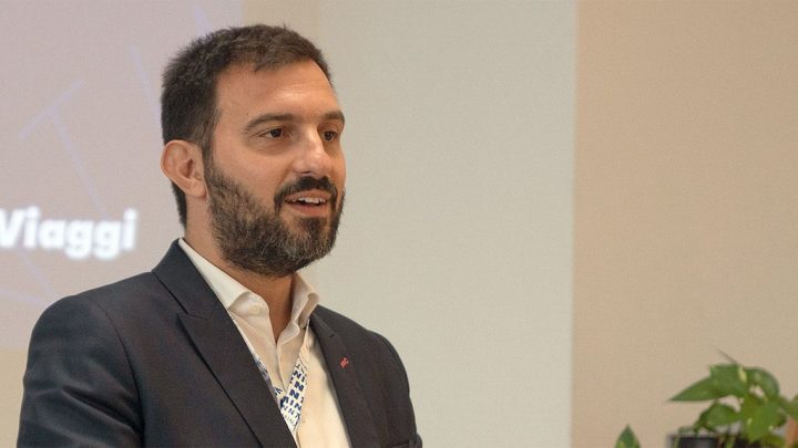 A man with dark hair and a beard in a suit jacket speaks about construction equipment tracking in front of a screen and plant.